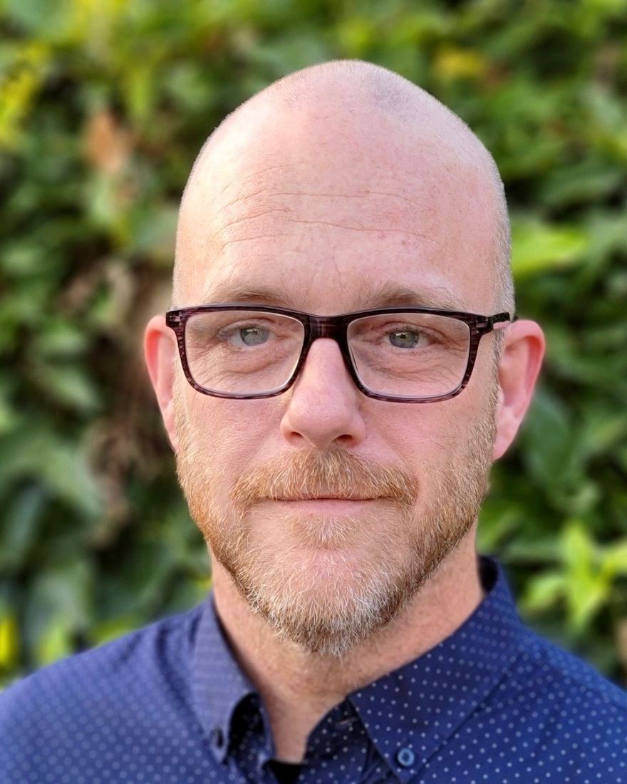 Headshot of a bearded man in dark-rimmed glasses and a navy patterned button-down shirt, photographed outdoors against green foliage.
