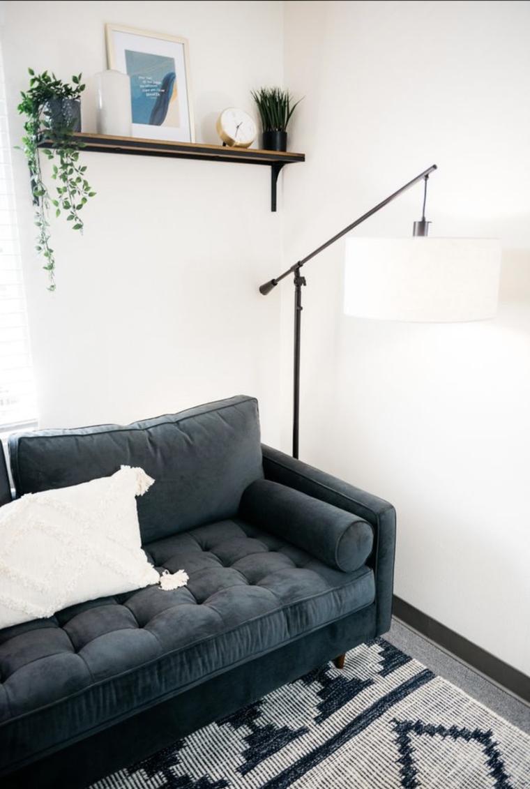 A consulting room corner: a deep blue-grey tufted sofa with a cream cushion, a black arc floor lamp, and a wooden shelf holding plants, a framed print and a brass clock against a white wall.