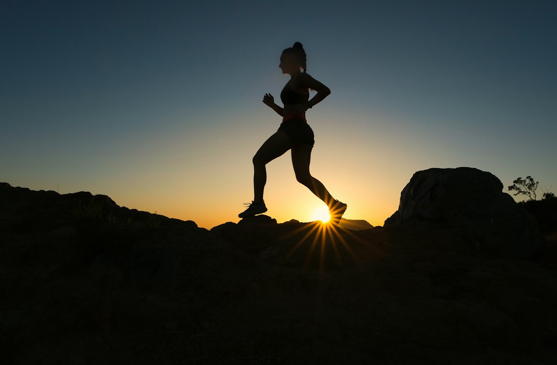A runner in silhouette on a rocky trail against a low sun.